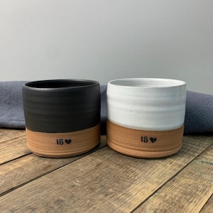 May include: Two handmade ceramic mugs. One is black, the other white, both with a natural clay base. Each mug has the number "18" and a heart symbol. The mugs are on a wooden surface.