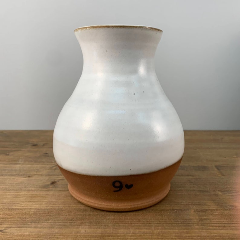 May include: White ceramic vase with a brown base. The vase has a rounded body and a wide opening. The base is marked with the number "9" and a heart.