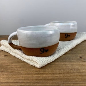 May include: Two white ceramic mugs with brown bottoms. Both mugs have a black "9" with a heart symbol on the bottom. The mugs are sitting on a white crocheted surface.