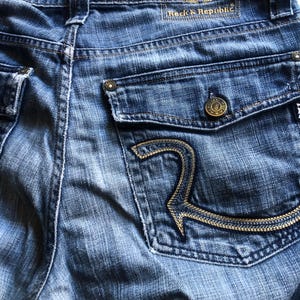 May include: Close-up of blue denim jeans with a back pocket featuring a gold embroidered "R" design. The jeans have a button closure and a "Rock & Republic" label with a winged logo.