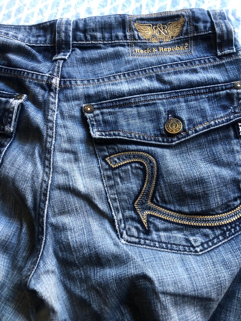 May include: Close-up of blue denim jeans with a back pocket featuring a gold embroidered "R" design. The jeans have a button closure and a "Rock & Republic" label with a winged logo.