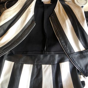 以下が含まれることがあります： 白と黒のストライプ柄のレザージャケットと、お揃いのスカート。 ジャケットはジッパー開閉で、白いストライプが入っています。 スカートは縦の白いストライプです。 この衣装は革製です。