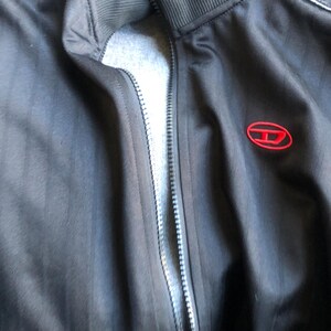 May include: Black track jacket with a full zipper and a red logo on the chest. The jacket has a ribbed collar and denim accents on the shoulders. The jacket is made of a smooth, dark fabric.