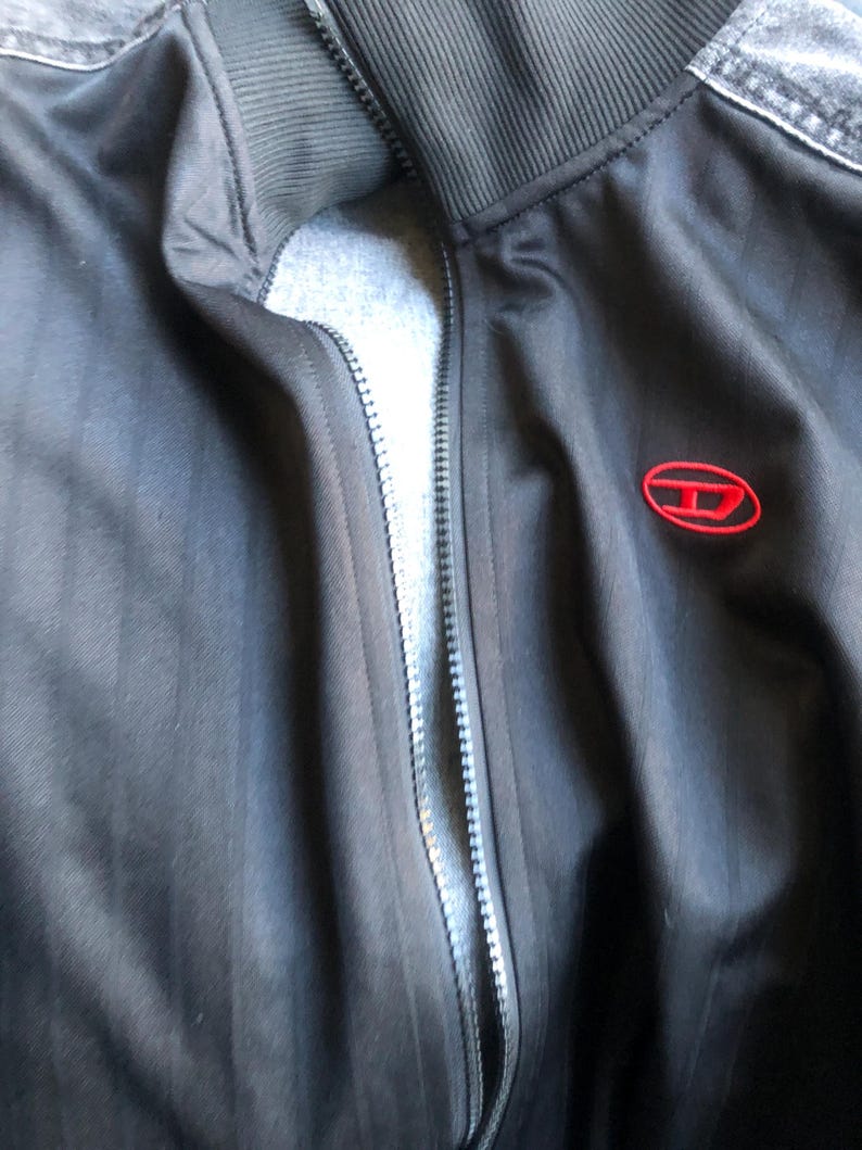 May include: Black track jacket with a full zipper and a red logo on the chest. The jacket has a ribbed collar and denim accents on the shoulders. The jacket is made of a smooth, dark fabric.