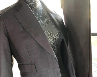 Puede incluir: Un blazer gris sobre un maniquí. El blazer tiene solapas con muescas, dos bolsillos delanteros y un corte entallado. La tela parece ser un material tejido. El cuello del maniquí está cubierto con un diseño de encaje blanco.