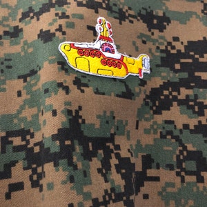 Peut inclure: Un écusson jaune en forme de sous-marin avec des détails rouges et blancs, cousu sur un fond de tissu camouflage.