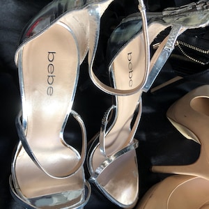 Puede incluir: Sandalias plateadas de tac&oacute;n alto con finas correas y la marca "bebe" visible en la plantilla. Las sandalias tienen un acabado met&aacute;lico brillante y un tac&oacute;n de aguja. Tambi&eacute;n se ven tacones color nude.