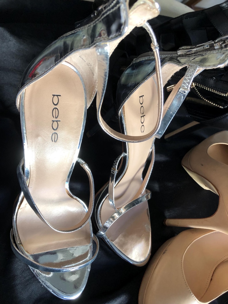 Puede incluir: Sandalias plateadas de tac&oacute;n alto con finas correas y la marca "bebe" visible en la plantilla. Las sandalias tienen un acabado met&aacute;lico brillante y un tac&oacute;n de aguja. Tambi&eacute;n se ven tacones color nude.