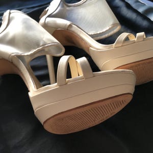 Puede incluir: Zapatos de tac&oacute;n alto con plataforma de color marfil con tacones de aguja. Los zapatos tienen m&uacute;ltiples correas y un detalle de malla en la parte superior. Las suelas son marrones. Los zapatos est&aacute;n sobre un fondo oscuro.