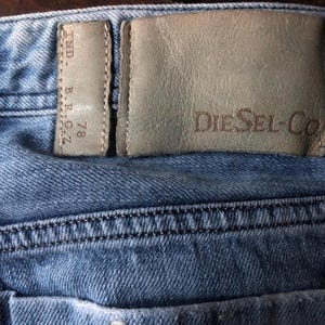 May include: Close-up of blue denim jeans with a leather patch. The patch is embossed with "Diesel-Co" and a smaller tag with "IND. B.R.G.Z. 78". The jeans have visible stitching and a faded appearance.