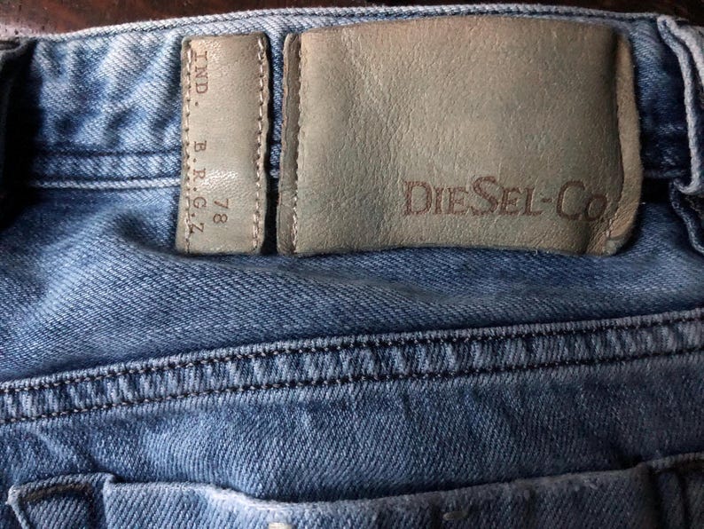 May include: Close-up of blue denim jeans with a leather patch. The patch is embossed with "Diesel-Co" and a smaller tag with "IND. B.R.G.Z. 78". The jeans have visible stitching and a faded appearance.