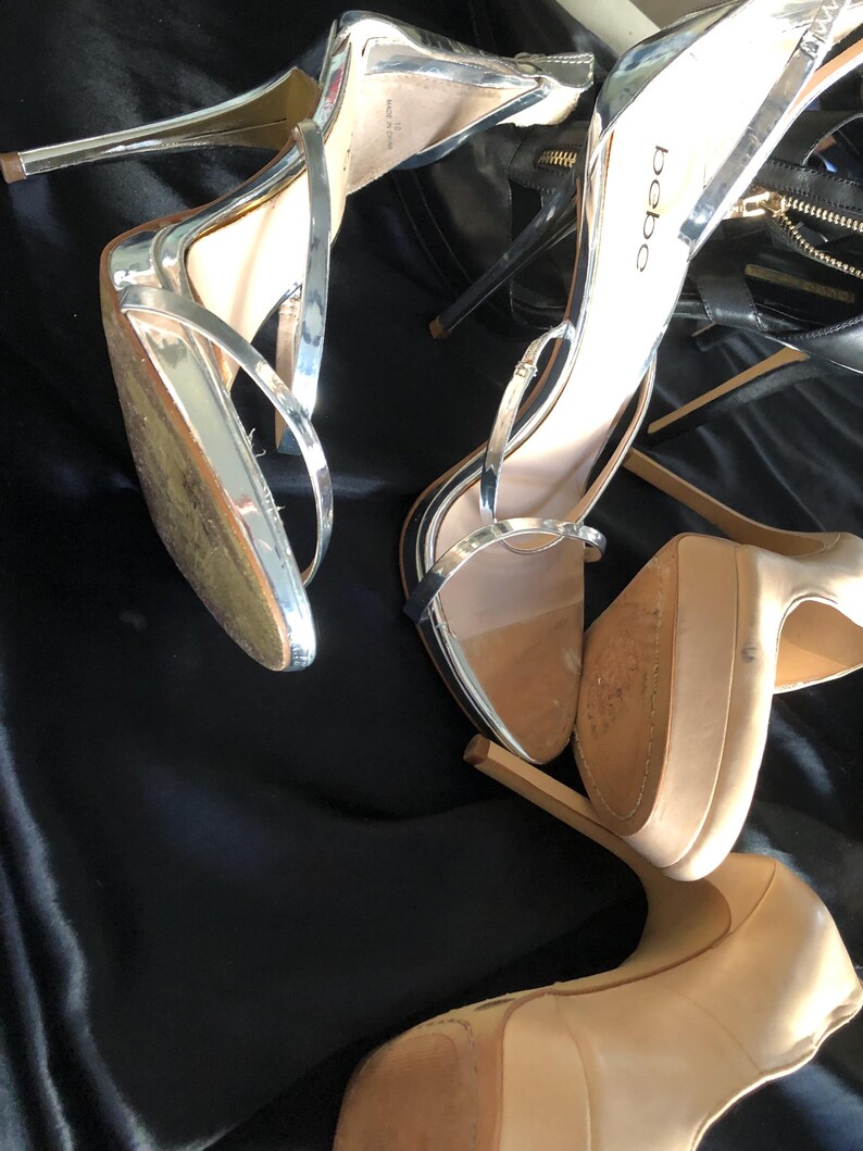 Puede incluir: Sandalias plateadas de tac&oacute;n alto con finas correas y tacones de aguja. Las sandalias tienen un brillo met&aacute;lico y est&aacute;n sobre una superficie negra. El nombre de la marca "bebe" es visible en uno de los zapatos.