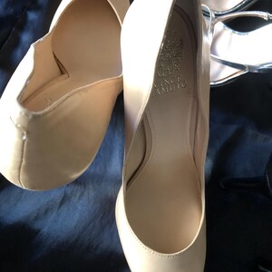 Puede incluir: Un par de tacones altos beige de Vince Camuto. Los zapatos tienen una punta redondeada y un tac&oacute;n medio. El interior del zapato es de color beige claro, y el nombre de la marca es visible dentro de uno de los zapatos. Los zapatos est&aacute;n sobre una superficie azul oscuro.