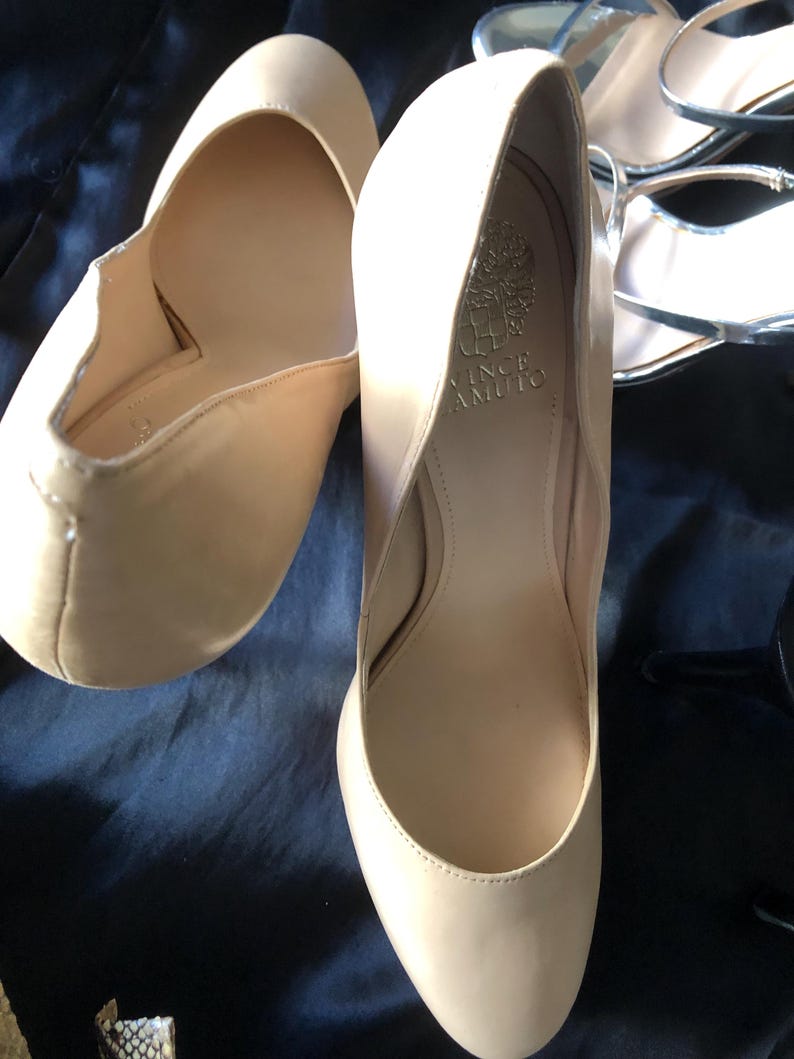 Puede incluir: Un par de tacones altos beige de Vince Camuto. Los zapatos tienen una punta redondeada y un tac&oacute;n medio. El interior del zapato es de color beige claro, y el nombre de la marca es visible dentro de uno de los zapatos. Los zapatos est&aacute;n sobre una superficie azul oscuro.