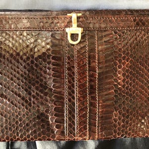 Pode incluir: Uma clutch de pele de cobra castanha com um fecho dourado. A clutch retangular apresenta um padrão texturizado e detalhes de costura verticais. O fecho é um design simples e elegante.