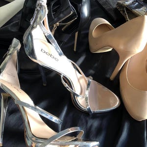 Puede incluir: Una colecci&oacute;n de zapatos de mujer, que incluye tacones plateados con tiras, zapatos de sal&oacute;n nude y botines negros. Los tacones plateados tienen tac&oacute;n de aguja y la marca "bebe" es visible. Los zapatos est&aacute;n colocados sobre una superficie negra.