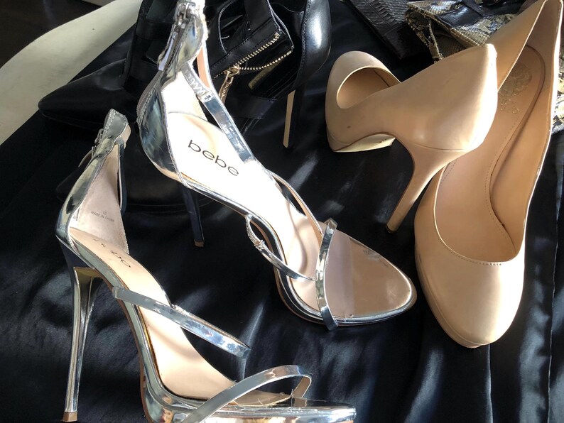 Puede incluir: Una colecci&oacute;n de zapatos de mujer, que incluye tacones plateados con tiras, zapatos de sal&oacute;n nude y botines negros. Los tacones plateados tienen tac&oacute;n de aguja y la marca "bebe" es visible. Los zapatos est&aacute;n colocados sobre una superficie negra.