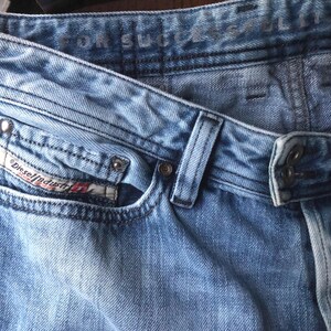 May include: Close-up of light blue denim jeans. The jeans feature a classic five-pocket design, belt loops, and a button and zipper closure. The Diesel Industry logo is visible on the front pocket. The jeans have a faded, worn look.