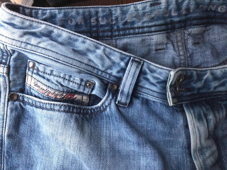 May include: Close-up of light blue denim jeans. The jeans feature a classic five-pocket design, belt loops, and a button and zipper closure. The Diesel Industry logo is visible on the front pocket. The jeans have a faded, worn look.