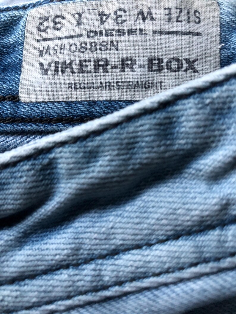 May include: Close-up of a light blue denim fabric with a label. The label reads "DIESEL" and "VIKER-R-BOX". The size is W34 L32. The jeans have a regular straight fit.