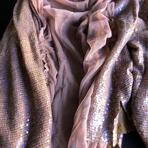 May include: A draped, open-front garment with a sheer, light pink mesh overlay and a sequined, bronze-colored outer layer. The garment has a flowing, layered design, with the sequins catching the light. The overall effect is a blend of textures and colors.