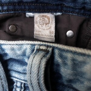 May include: Close-up of blue denim jeans with a faded wash. The interior label reads "SIZE 34" and "MADE IN TUNISIA". Two silver rivets and a dark brown interior lining are visible.