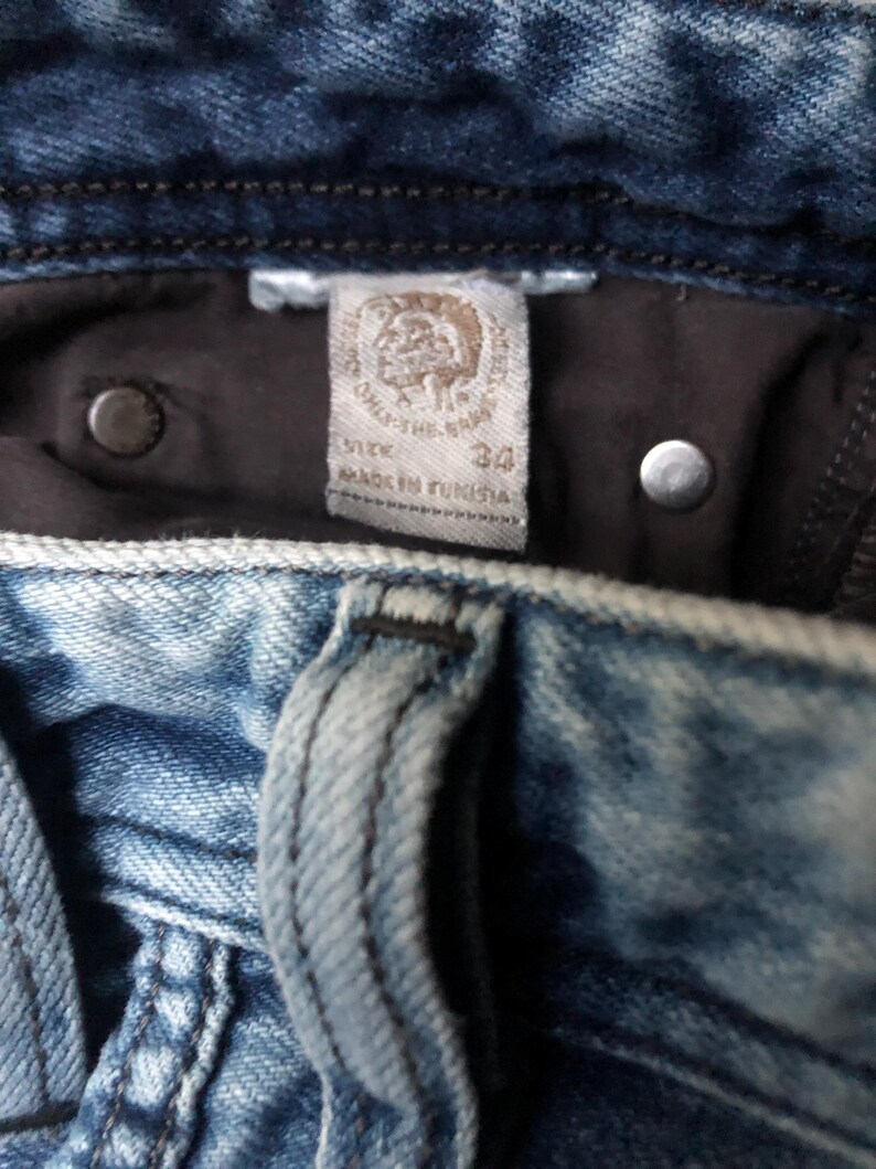 May include: Close-up of blue denim jeans with a faded wash. The interior label reads "SIZE 34" and "MADE IN TUNISIA". Two silver rivets and a dark brown interior lining are visible.