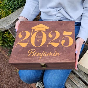 May include: Two wooden boxes with engraved graduation designs. The boxes are brown and natural wood. The top box has the text "Class of 2025" in gold lettering and the name "Benjamin" below. The bottom box has the text "Class of 2025" in brown lettering and the name "Benjamin" below.