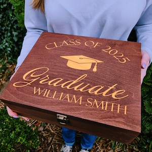 May include: A wooden box with a gold inscription that reads "Class of 2023 Graduate William Smith". The box has a gold graduation cap design.