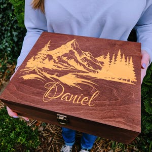 May include: A wooden box with a hinged lid, engraved with a mountain scene and the name "Daniel". The box is made of dark wood with a reddish brown stain and features a gold latch.