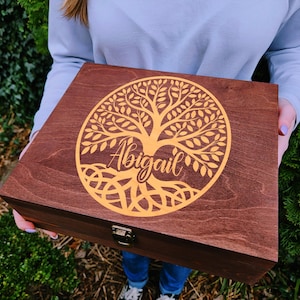 May include: A wooden box with a tree of life design and the name "Abigail" in a gold color. The box is rectangular with a dark brown finish and a metal clasp. The tree design is centered on the lid.