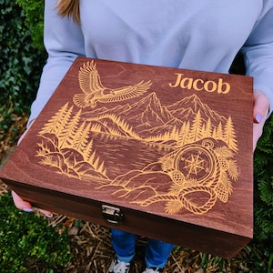 May include: A rectangular dark brown wooden box featuring a laser-engraved design on the lid. The engraving depicts a mountain landscape with a lake, pine trees, a flying eagle, a compass, and rope. The name Jacob is engraved in the top right corner.