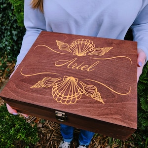 May include: A wooden box with a hinged lid, engraved with a seashell design and the name "Ariel". The box is a rich brown color and has a gold latch.