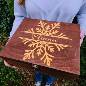 May include: A dark brown wooden box featuring a gold snowflake design and the name "Eliana". The box has a metal clasp and is held by a person. The box is likely for storage or gifting.