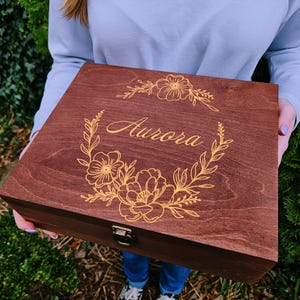 May include: A rectangular dark brown wooden box featuring a gold-coloured engraved floral wreath design on the lid. The name Aurora is centred within the wreath in a cursive script. The box includes a small metal latch on the front edge.