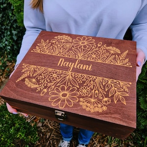 May include: A rectangular dark brown wooden box with a hinged lid featuring a laser-engraved floral pattern and the name Naylani centered in a horizontal band. The box includes a small metal latch on the front edge for secure closure.