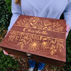 May include: A wooden box with a dark brown finish, featuring a floral design and the name "Charlotte" in gold lettering. The box is rectangular with a hinged lid and a metal clasp.