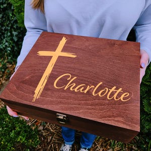 May include: A wooden box with a hinged lid, stained a dark brown. The lid features a gold-colored cross and the name "Charlotte" in a script font. The box is held by a person.