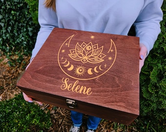 May include: A wooden box with a dark brown finish, featuring a gold celestial design of a lotus flower, crescent moons, and stars. The name "Selene" is inscribed in gold script below the design. The box has a metal clasp.