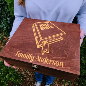 May include: A wooden box with a hinged lid, stained a rich brown. The lid features a gold illustration of an open book with the words "HOLY BIBLE" and "Family Anderson" in gold lettering. A metal clasp secures the box.
