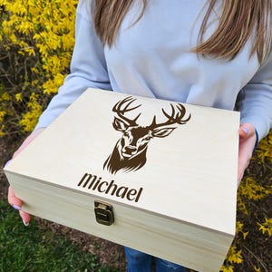 May include: A light-colored wooden box with a brown deer head design and the name "Michael" printed on the front. The box has a metal clasp and is being held in front of a yellow flowering bush.