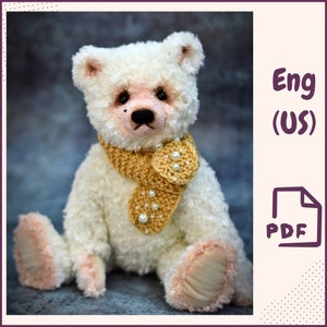 Puede incluir: Un oso de peluche blanco con una bufanda de crochet amarilla y blanca con detalles de perlas. El oso está sentado sobre una superficie gris texturizada. Eng (US) PDF.