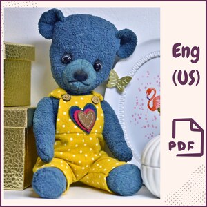 May include: A blue teddy bear wearing yellow overalls with white polka dots and a red, brown, and green heart applique. The bear is sitting on a white surface with a gold box and a white plate with pink flamingos in the background. The text "Eng (US)" and "PDF" are visible in the image.