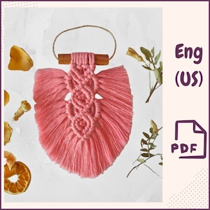 May include: A pink macrame wall hanging with a wooden dowel and fringe. The hanging is in the shape of a leaf and has a knot pattern.