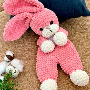 May include: A plush crochet bunny toy made of soft pink yarn with white accents on the paws, feet, and muzzle. The stuffed animal features long, floppy ears, small black bead eyes, and a stitched nose, crafted with a textured, chunky yarn finish.