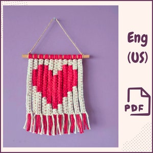May include: A macrame wall hanging featuring a red heart design against a white and cream background. The piece is suspended from a wooden dowel with a jute string and has a fringe at the bottom. The background is a solid purple.