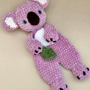 May include: A handmade crochet koala lovey crafted from soft, textured lavender yarn. The koala features a dark brown nose, white inner ears, a white belly patch, and a small green leaf held in its paws. It has black safety eyes with blue accents.