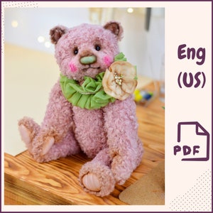 May include: A pink teddy bear with green and brown accents. The bear is wearing a green ruffled collar and a brown flower with a pearl center. The bear is sitting on a wooden surface.