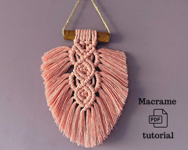 Peut inclure: Une suspension murale en macram&eacute; rose avec une baguette en bois en haut. La suspension a la forme d'un c&oelig;ur et a une frange autour des bords. Le texte "Tutoriel macram&eacute; PDF" est sous la suspension.