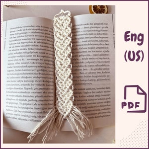 May include: A close-up of a white crocheted bookmark with a fringe. The bookmark is on top of an open book with text in a foreign language. The text on the book reads "Eng (US) PDF".